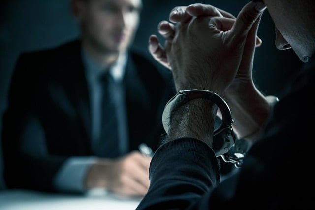 A close-up of a man's hands handcuffed together in an interrogation room with a man in the suit facing him in the background.